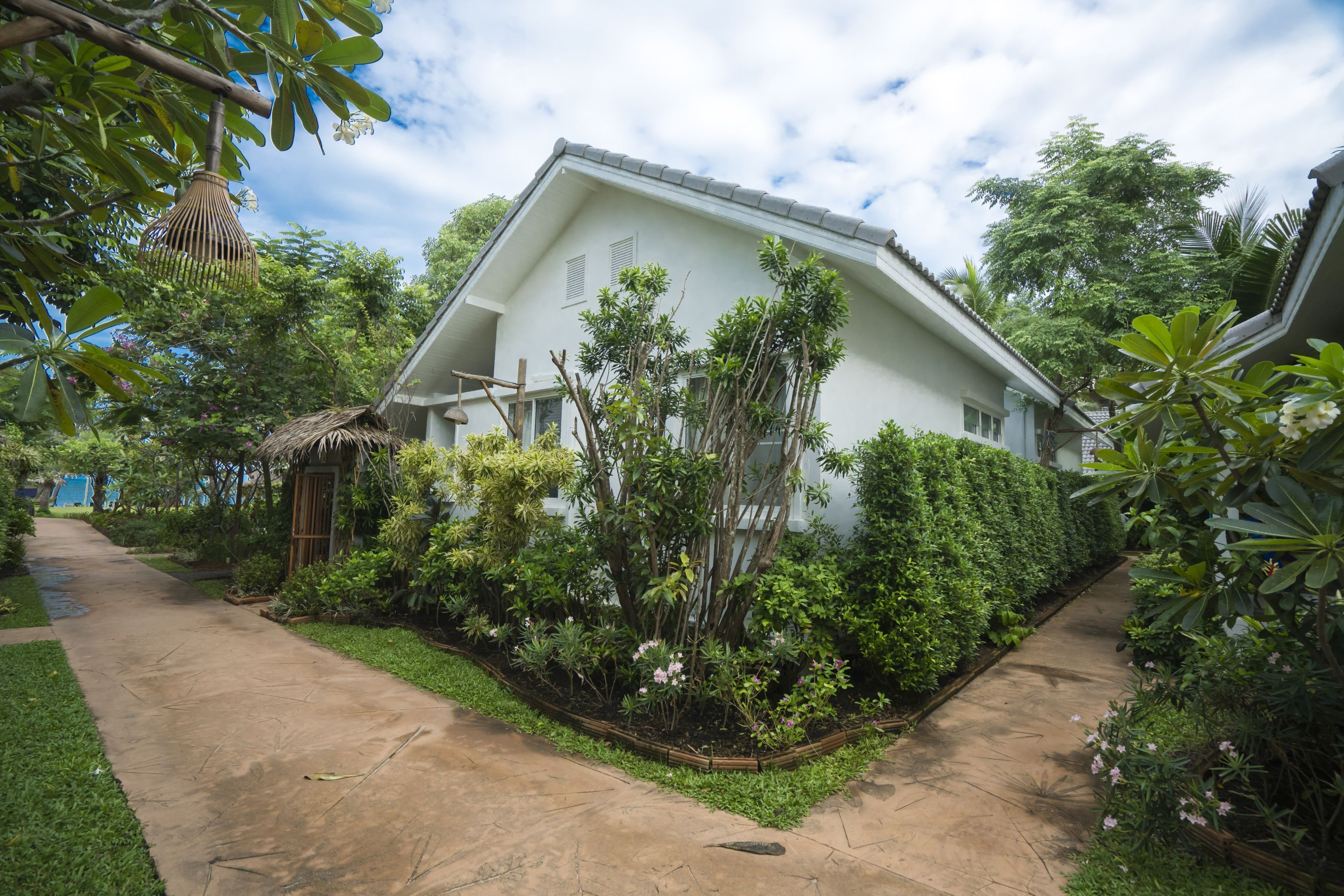 A white house with a sloped roof, surrounded by lush greenery and a concrete pathway.