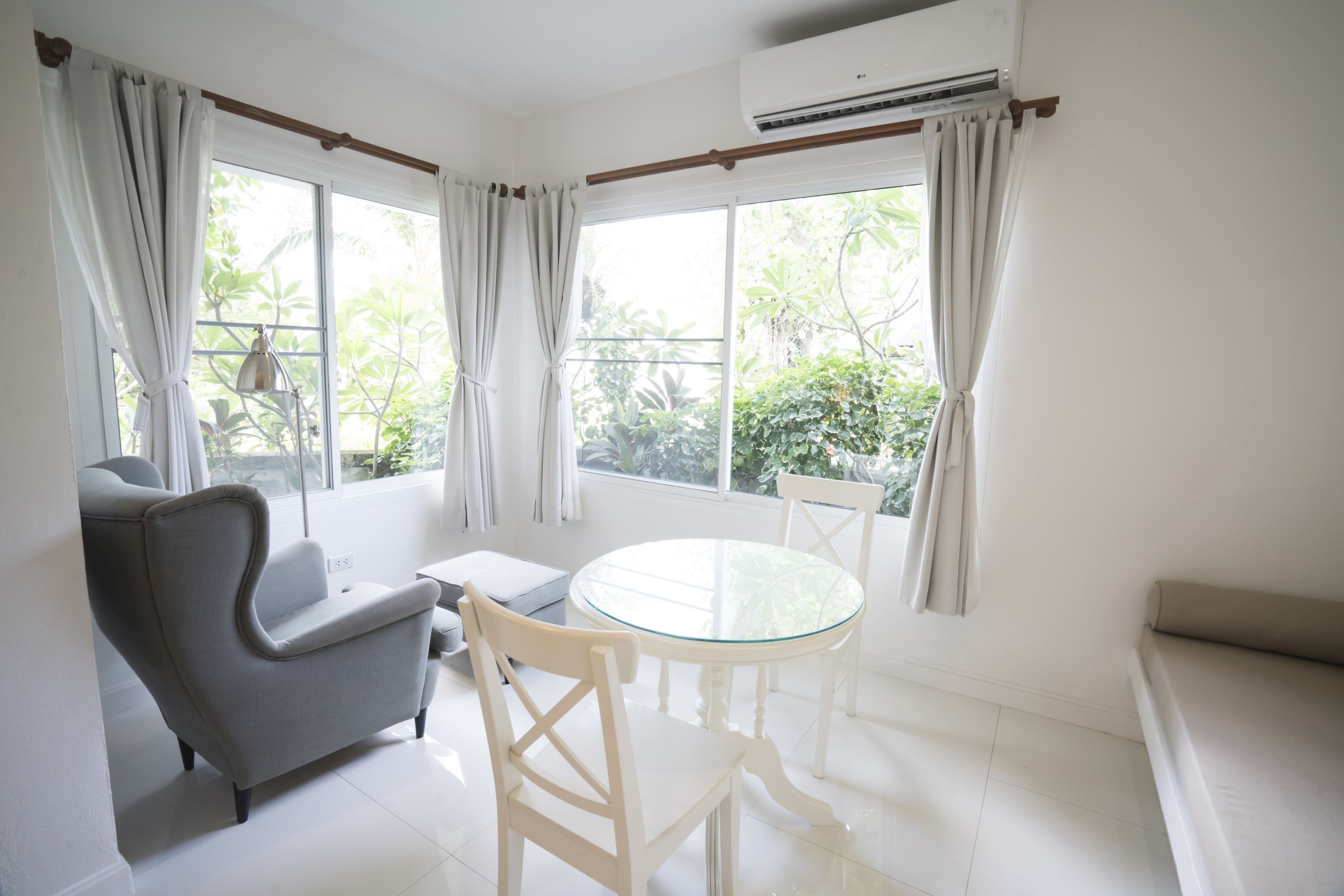 A room with a round glass table, two white chairs, a grey armchair, and a sofa. There are two windows with curtains, and an air conditioning unit above one window.