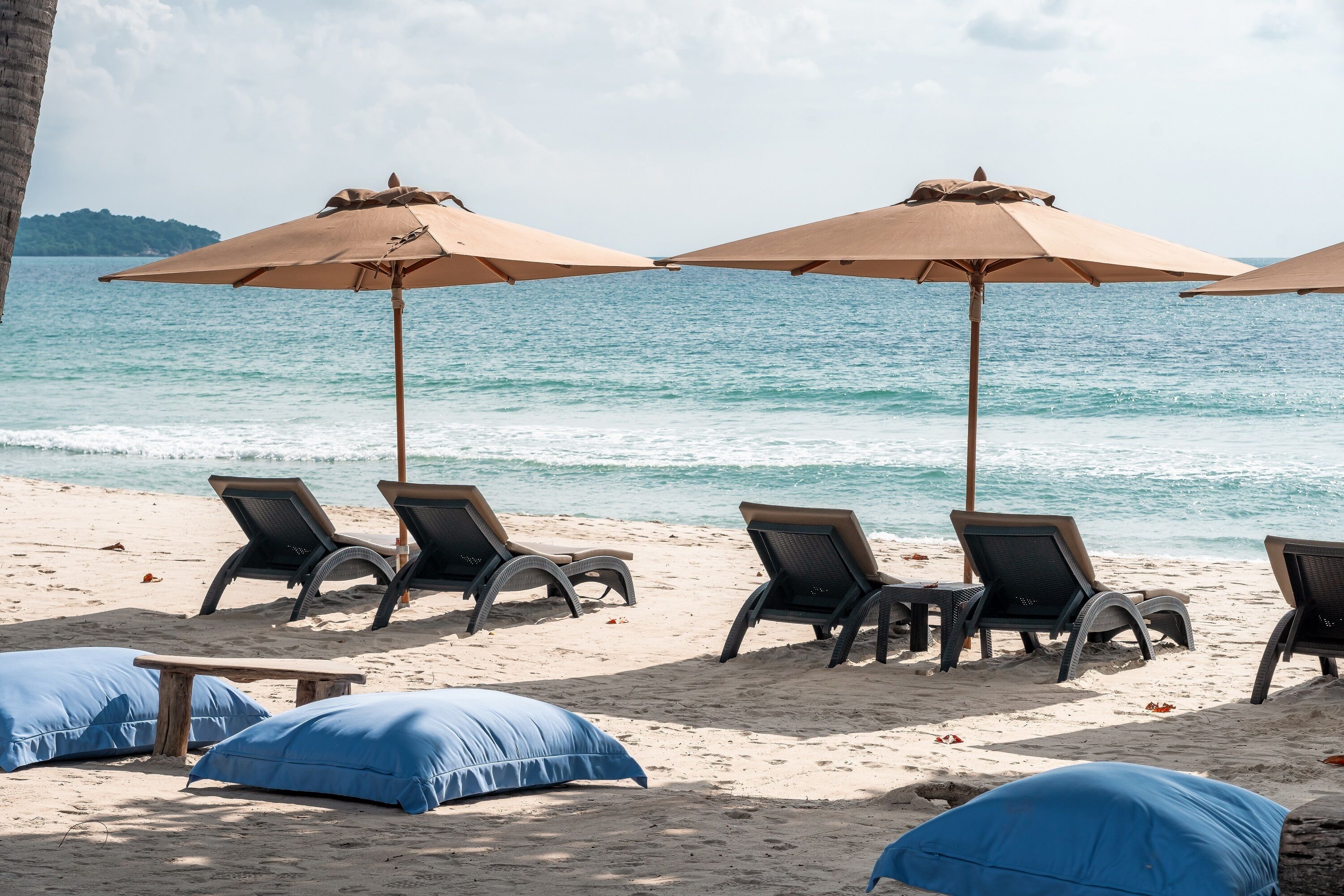 A beach with lounge chairs and umbrellas, a wooden table, and blue cushions.