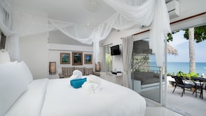 A large bed with white linens, a blue pillow, and white towels. There are framed pictures on the wall, a TV mounted above the bed, and a glass door leading to a balcony with a view of the ocean. - King's Garden Resort (Koh Samui)