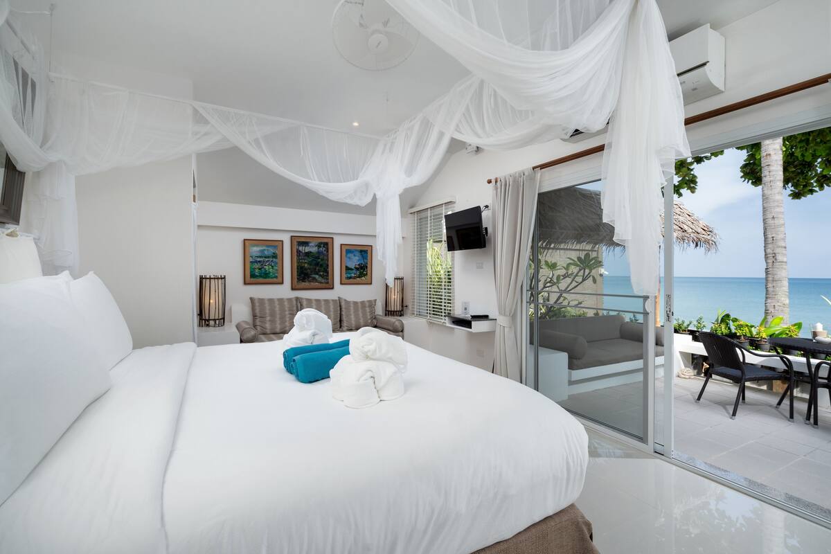 Ein großes Bett mit weißen Laken, einem blauen Kopfkissen und weißen Handtüchern. An der Wand befinden sich gerahmte Bilder, ein Fernseher ist über dem Bett montiert und eine Glastür führt zu einem Balkon mit Meerblick.