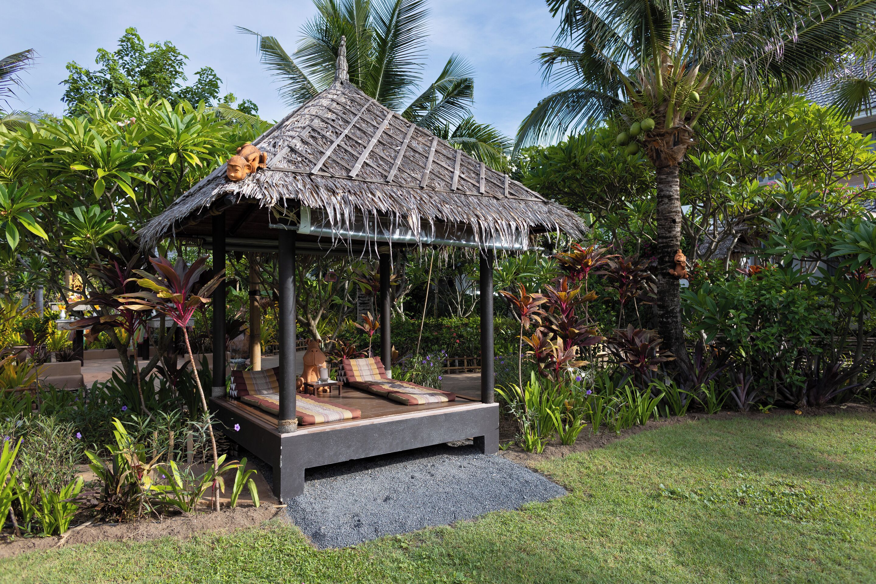 A thatched-roof gazebo with a bed and a small table, surrounded by lush tropical plants.