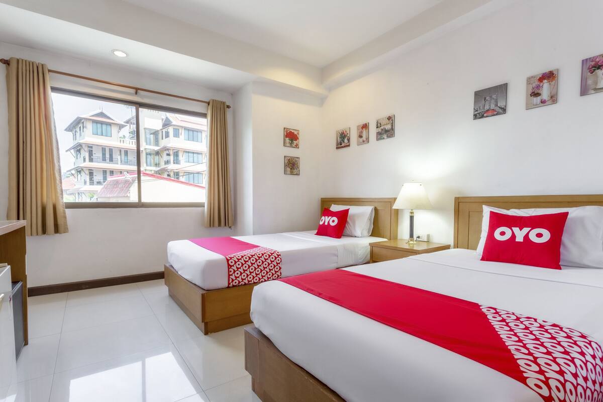 A hotel room with two beds, each with a red 'OYO' pillow, a bedside table with a lamp, and a window with curtains.