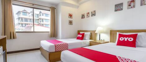 A hotel room with two beds, each with a red 'OYO' pillow, a bedside table with a lamp, and a window with curtains.