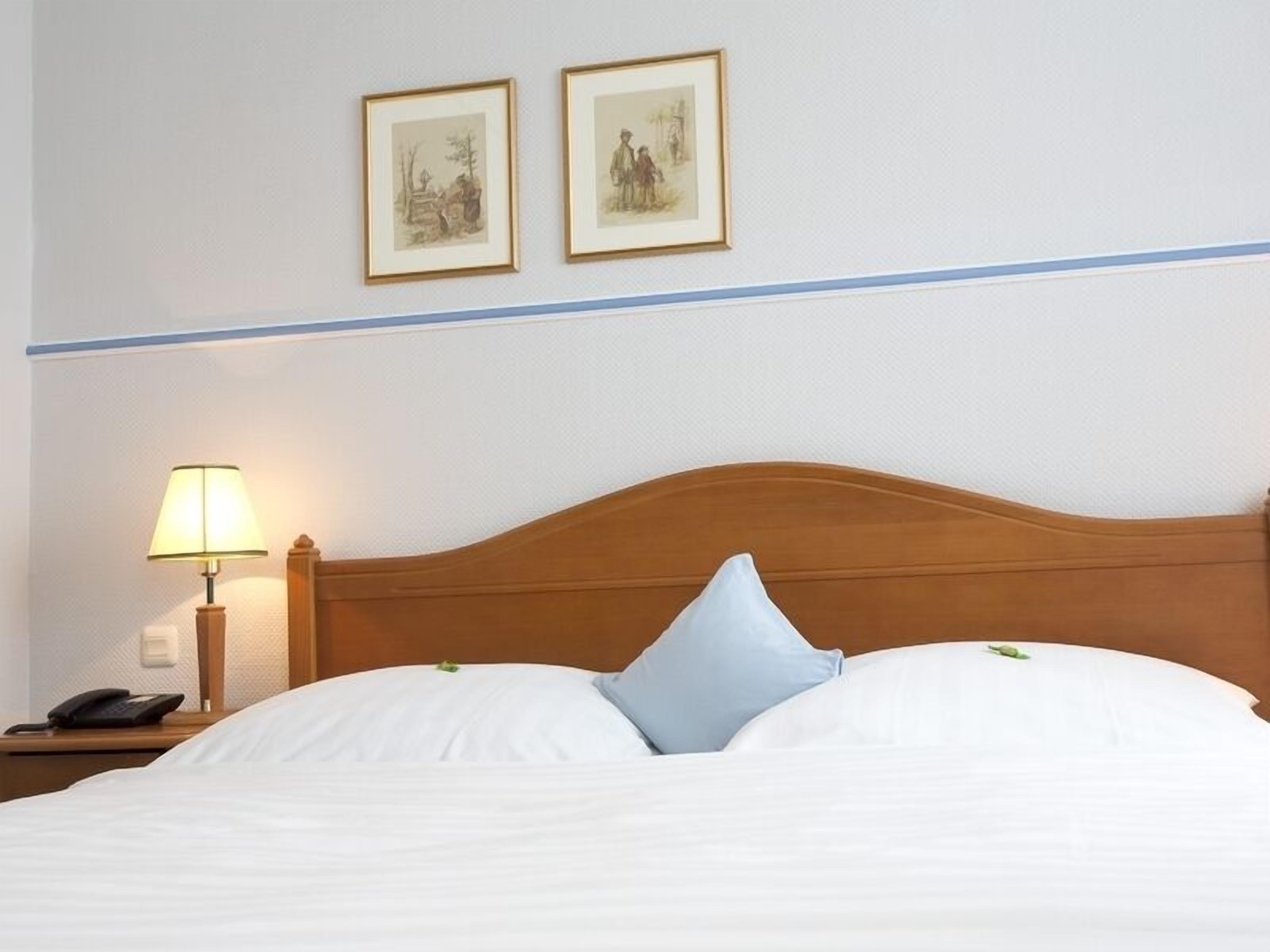 Una cama bien hecha con sábanas blancas y cabecera de madera, una lámpara de noche y dos cuadros en la pared.