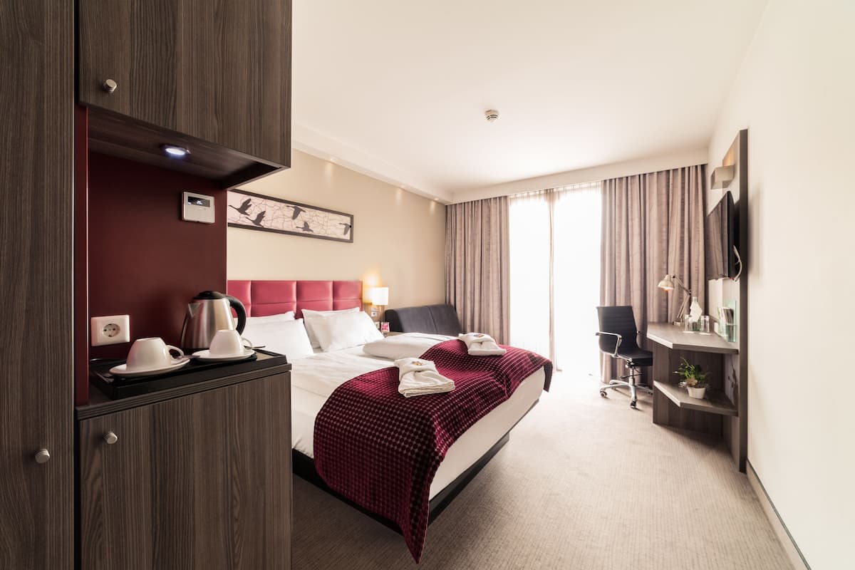 A hotel room with a bed, bedside table, lamp, and a view of the outdoors.