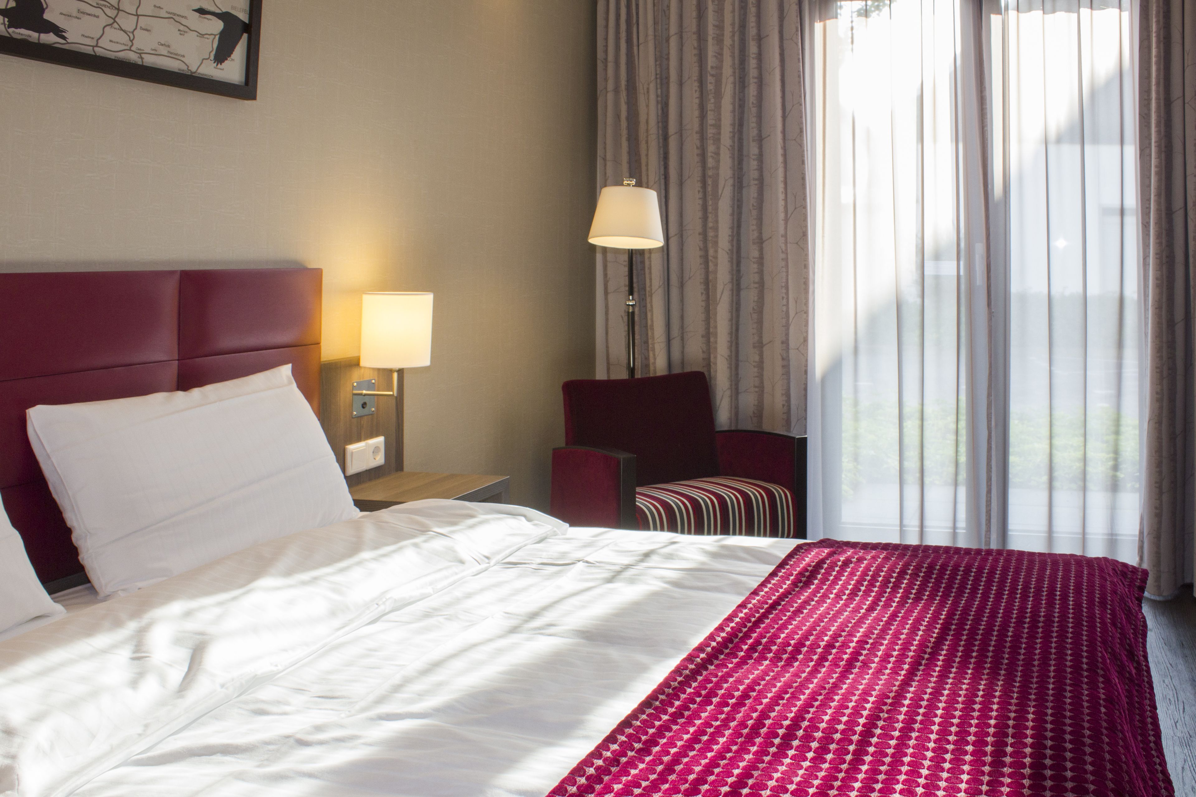 Ein Hotelzimmer mit einem großen Bett, einem roten Sessel, einem Nachttisch mit Lampe und Blick ins Freie durch ein Fenster mit Vorhängen.