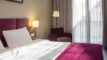 Ein Hotelzimmer mit einem großen Bett, einem roten Sessel, einem Nachttisch mit Lampe und Blick ins Freie durch ein Fenster mit Vorhängen.