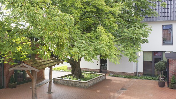 A courtyard with a large tree, a wooden bench, and a brick building. - Hotel zur Krone (Gescher)