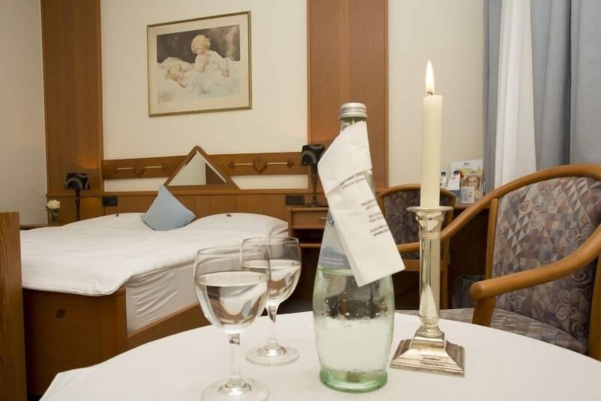A hotel room with a neatly made bed, a table with a bottle and glasses, a candle, and a painting on the wall.