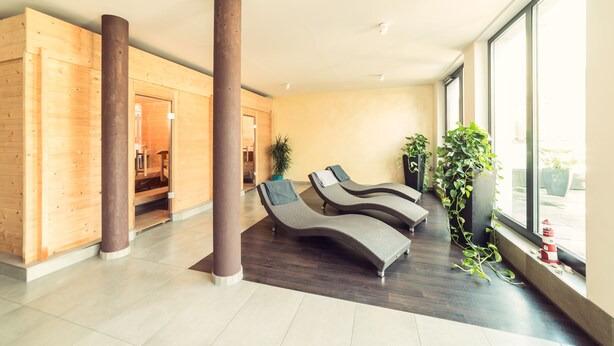 A modern spa area with wooden walls, two lounge chairs, a large potted plant, and a view of the outdoors through large windows. - Hotel zur Krone (Gescher)