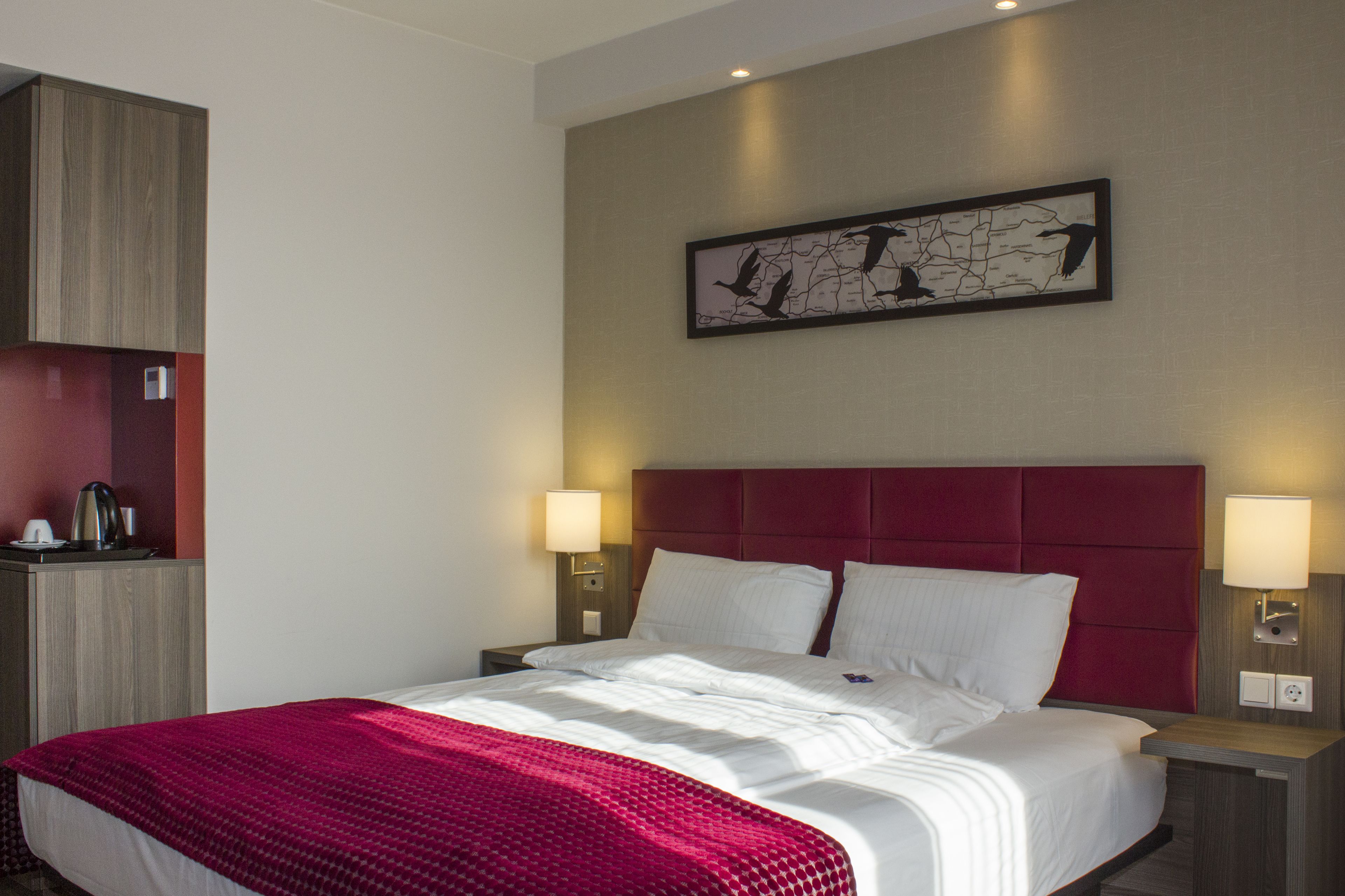 Une chambre d’hôtel avec un grand lit, deux tables de chevet, une tête de lit rouge et une œuvre d’art représentant des oiseaux, accrochée au mur.