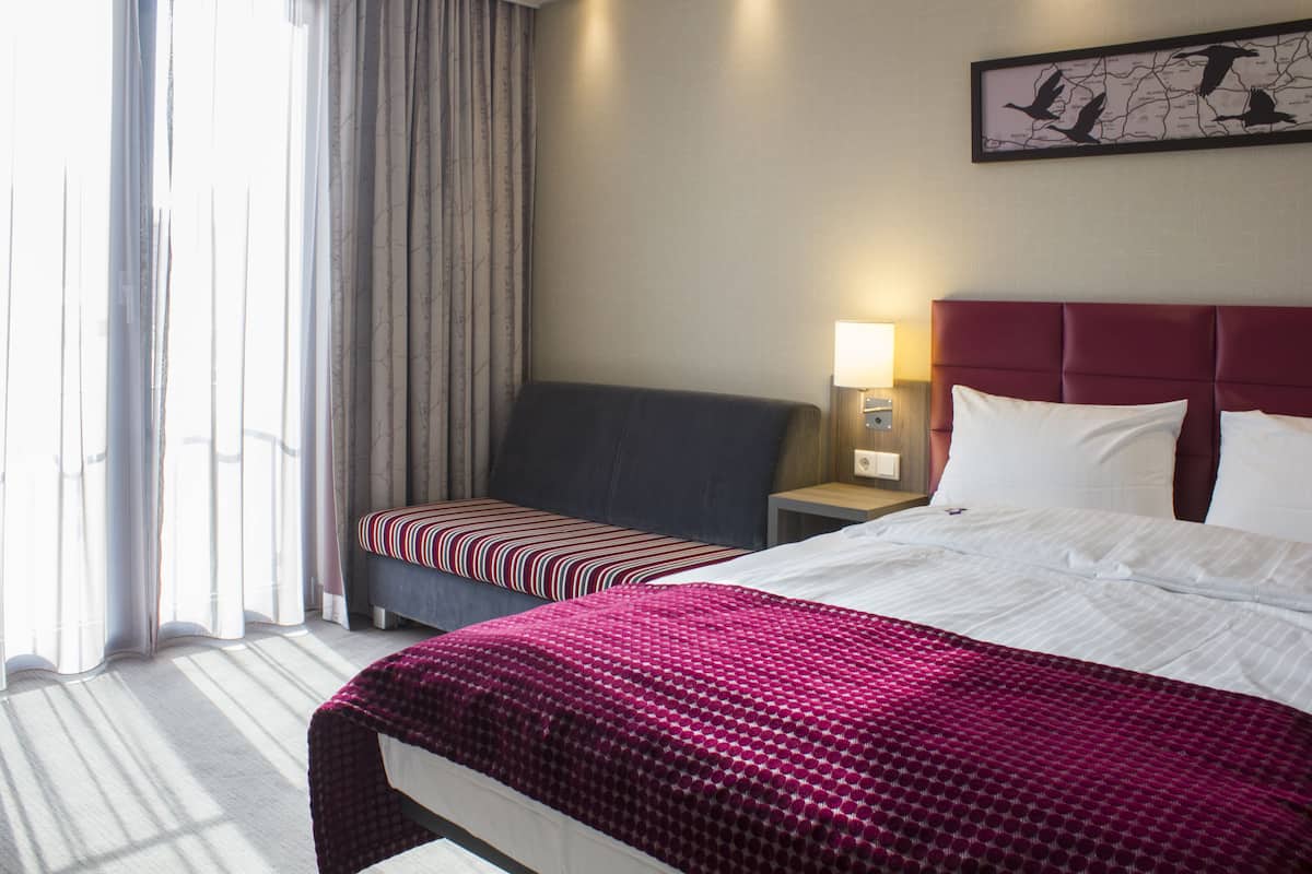 A hotel room with a large bed, a smaller bed, a nightstand with a lamp, a window with curtains, and a wall-mounted artwork.