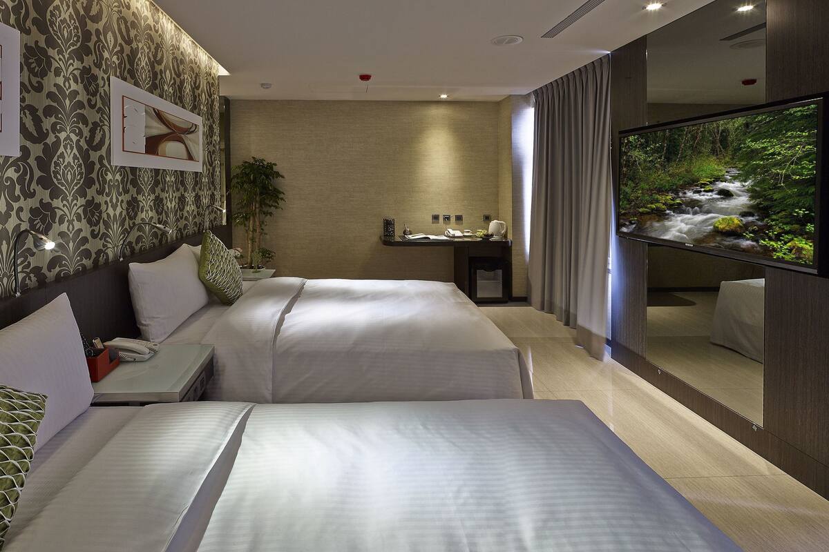 Een hotelkamer met twee bedden, een bureau, een televisie en een badkamer met spiegel en gordijnen.