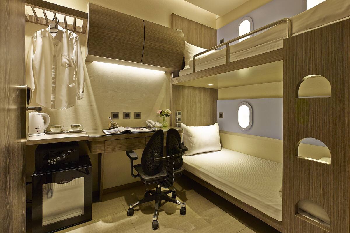 Een kleine hotelkamer met stapelbedden, een bureau met een computer, een stoel, een koffiezetapparaat en een bijzettafeltje met een vaas.