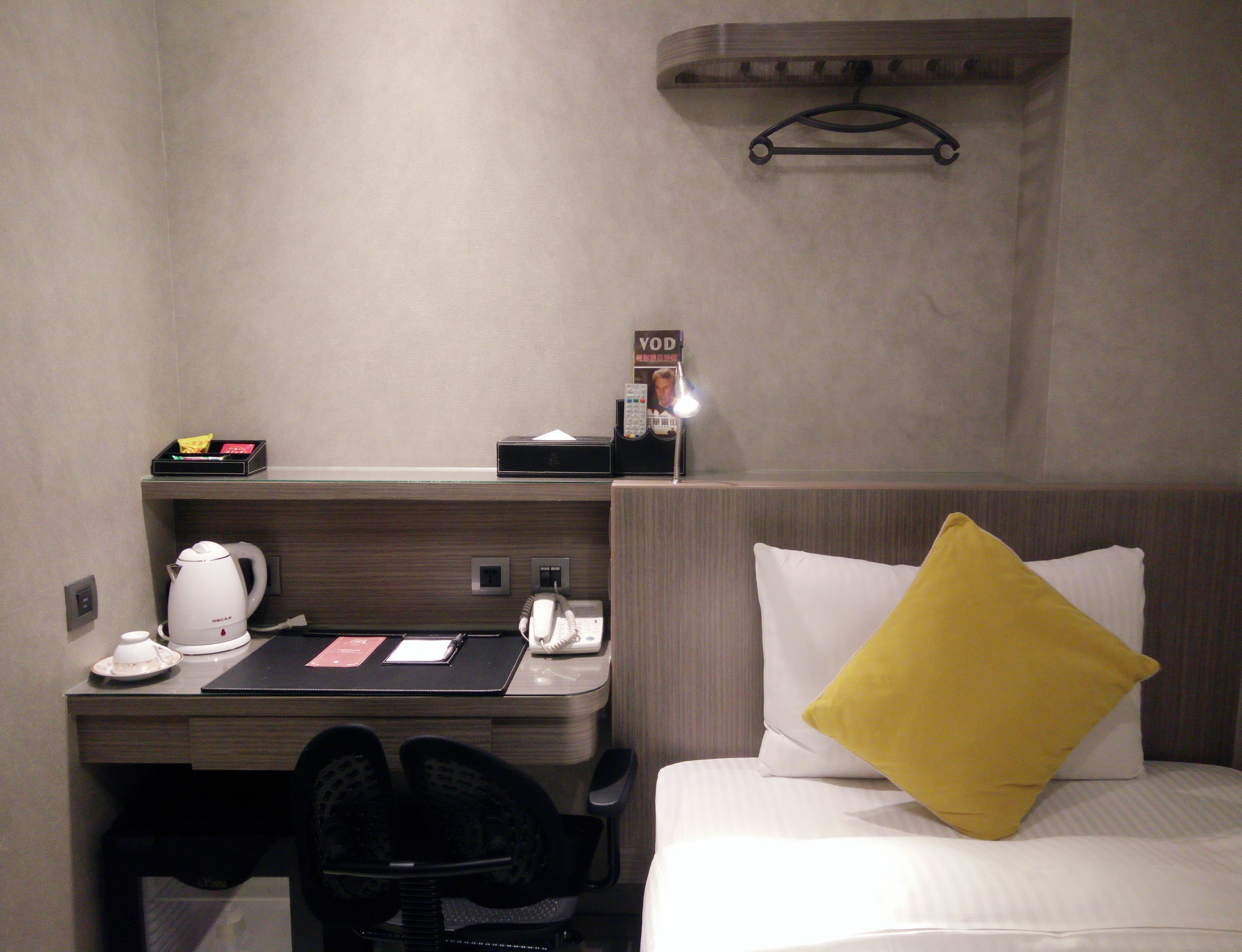 A hotel room with a desk, a telephone, a kettle, and a bed with a yellow pillow.