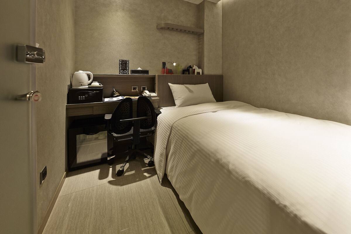 Een kleine hotelkamer met een enkel bed, een bureau met stoel, een koffiezetapparaat en een telefoon.