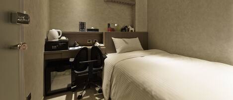 A compact hotel room with a single bed, a desk with a chair, a coffee maker, and a telephone.