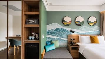 A hotel room with a bed, bedside table, desk, chair, and a wall with artwork.