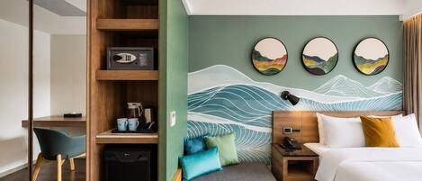 A hotel room with a bed, bedside table, desk, chair, and a wall with artwork.