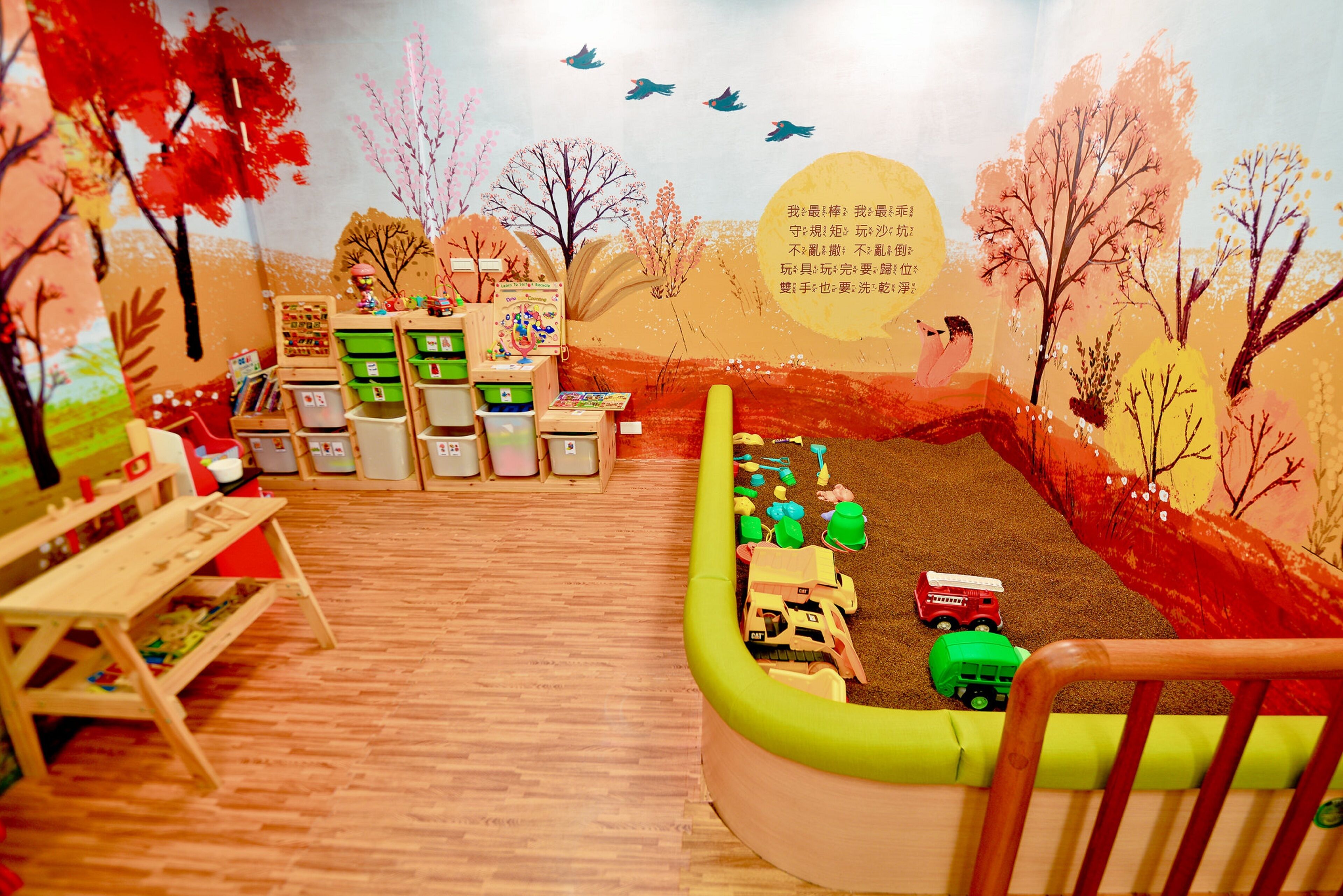 A children's playroom with wooden flooring, colorful play structures, and a mural of a landscape with trees and birds.