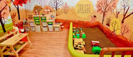 A children's playroom with wooden flooring, colorful play structures, and a mural of a landscape with trees and birds.