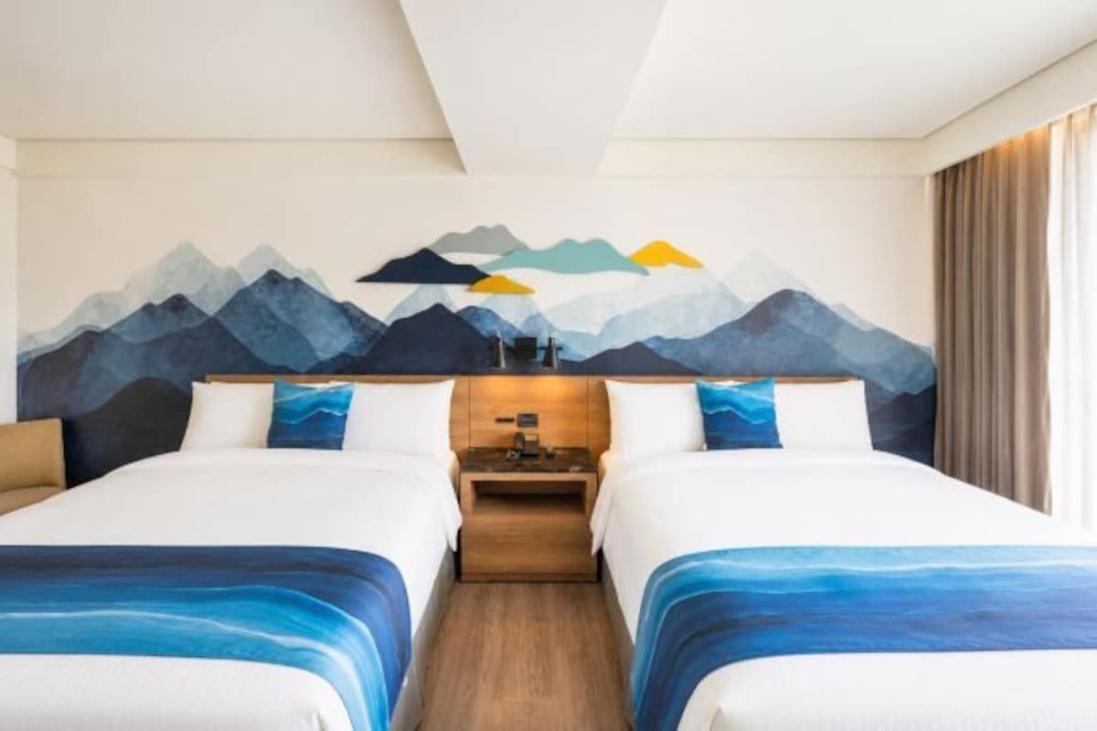Habitación de hotel con dos camas, un cuadro de montañas, una mesita de noche y una ventana con cortinas.