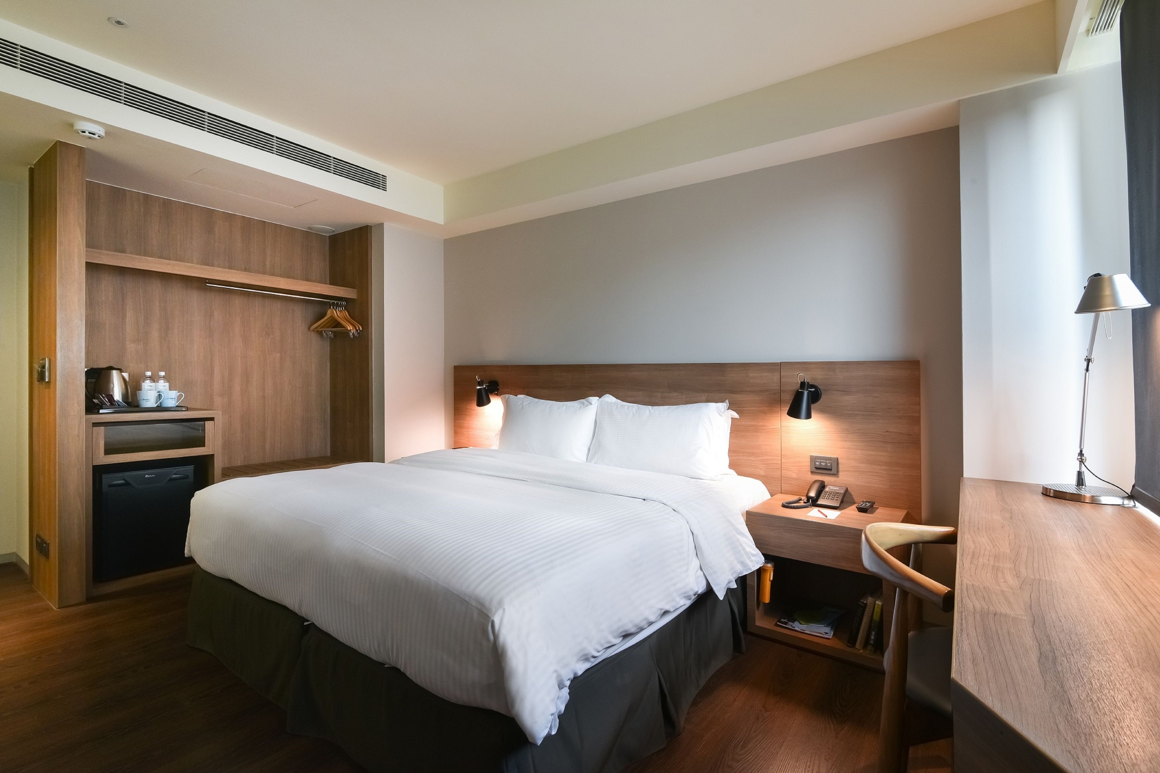 A modern hotel room with a large bed, wooden headboard, bedside tables with lamps, a built-in wardrobe, and a small desk with a lamp.