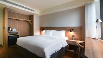 A modern hotel room with a large bed, wooden headboard, bedside tables with lamps, a built-in wardrobe, and a small desk with a lamp.