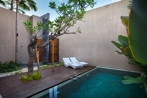 Un área de piscina moderna al aire libre con una terraza de madera, dos sillones blancos y un árbol pequeño.