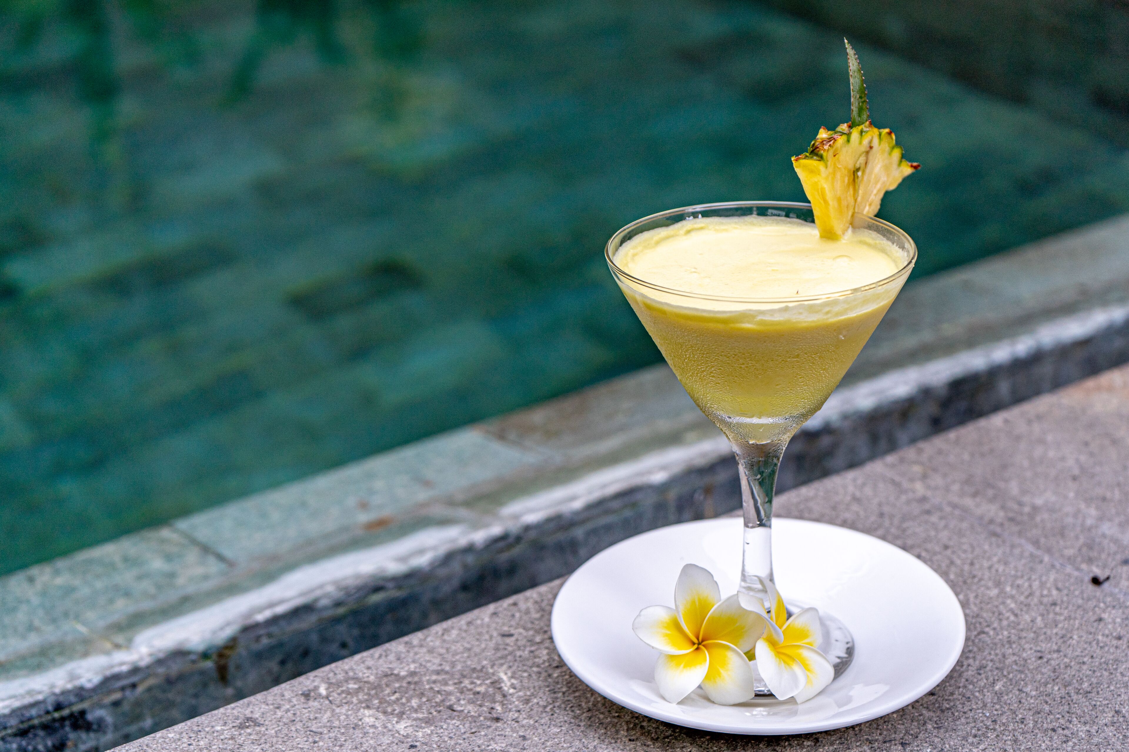 Een cocktail in een martini-glas met garnering op een wit bord, geplaatst op een stenen rand bij een zwembad.