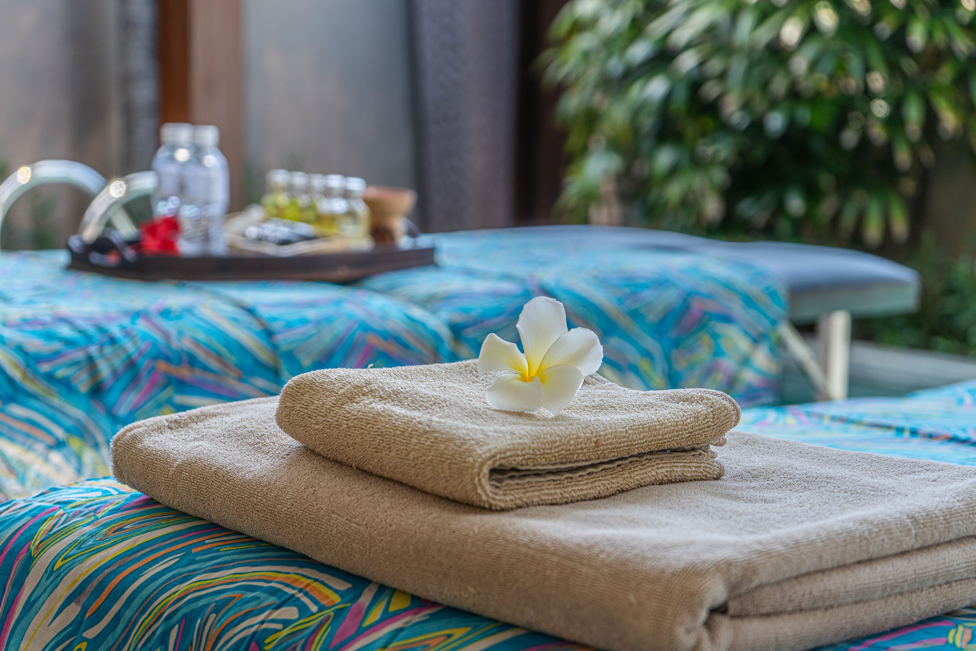 A neatly folded beige towel with a white flower on a bed with a colorful, abstract-patterned bedspread.