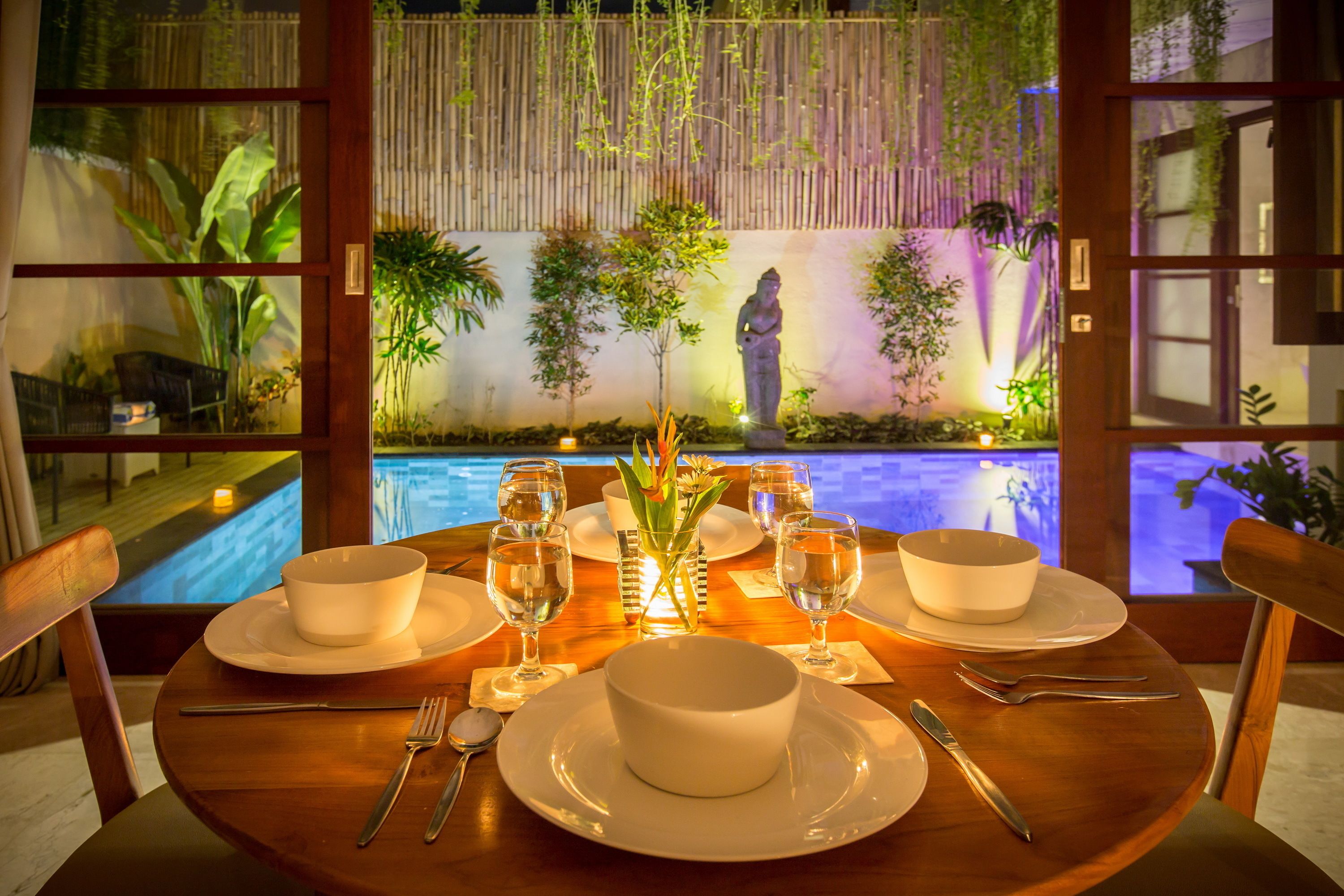 A dining table set for two with white dishes and cutlery, a vase with flowers, and a view of a swimming pool and bamboo plants through a wooden door.