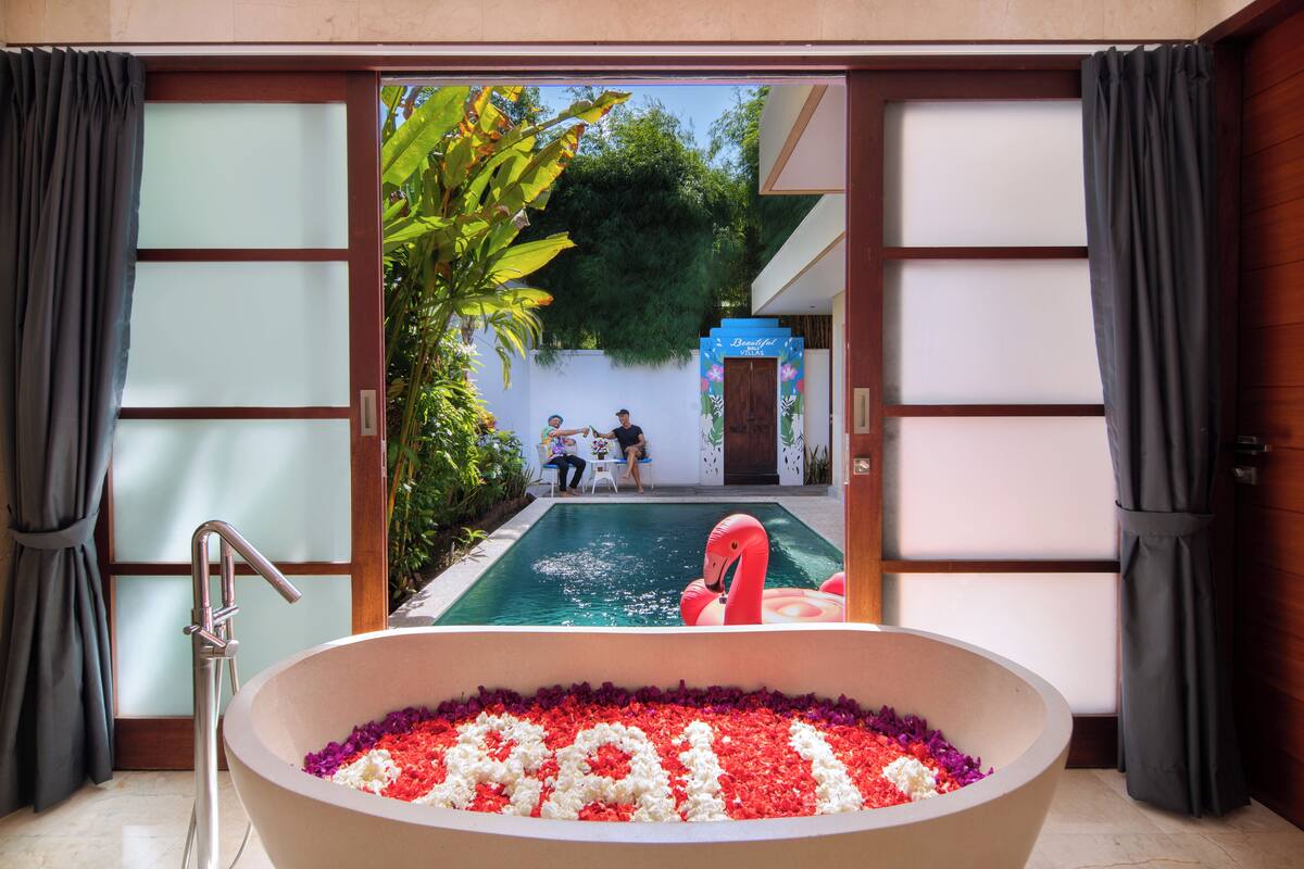 A large bathtub filled with water and flower petals, with a view of a swimming pool and a flamingo float outside.