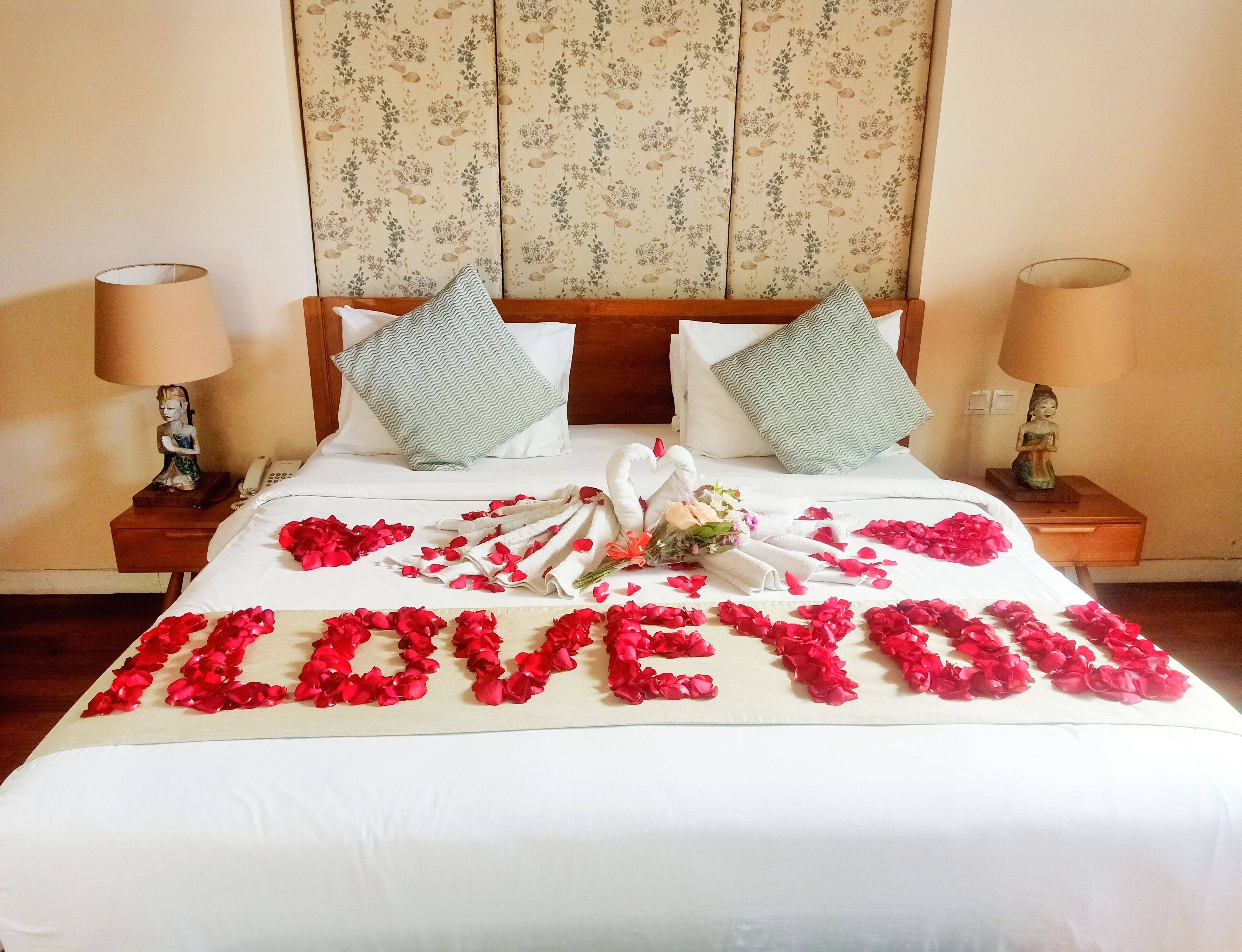 Une chambre d’hôtel avec un lit décoré de pétales de rose formant les mots « JE T’AIME » et un bouquet de fleurs.