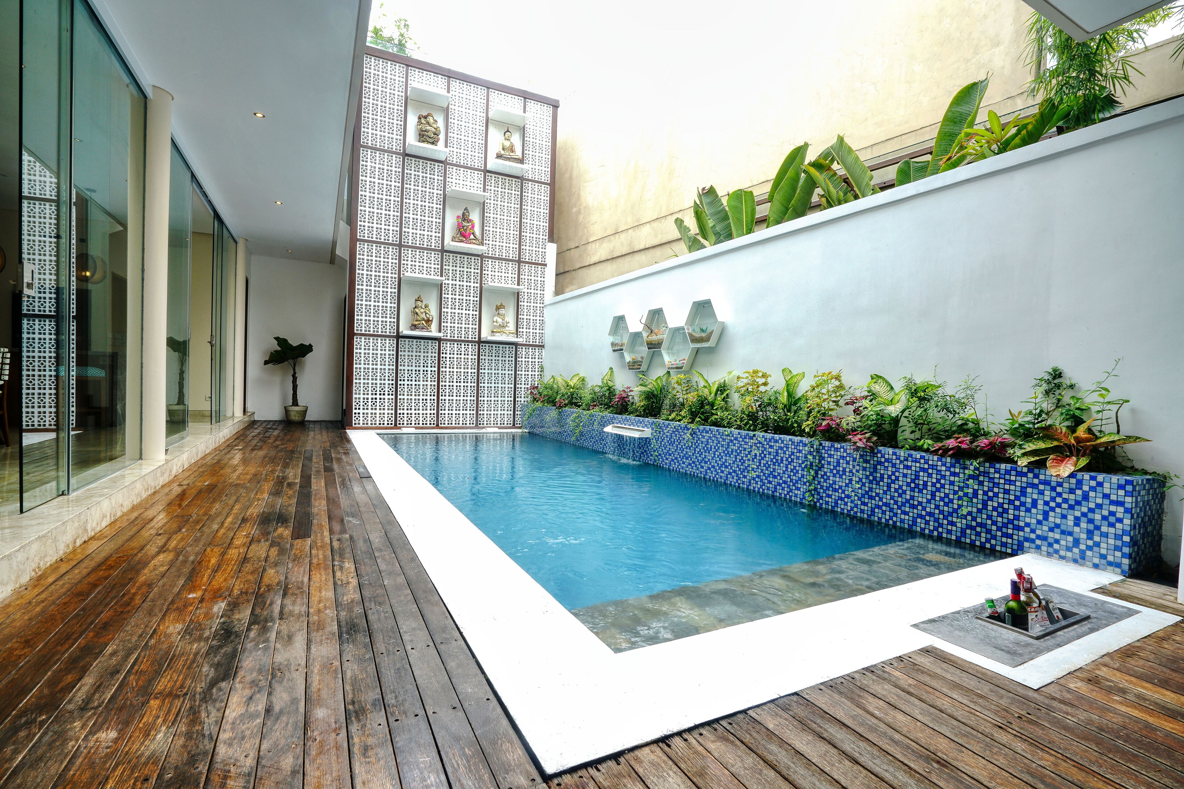 A modern pool area with a wooden deck, a blue mosaic wall, and various plants.