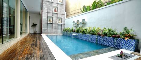 A modern pool area with a wooden deck, a blue mosaic wall, and various plants.
