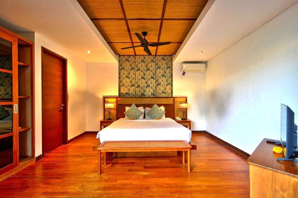 A hotel room with a wooden ceiling, a bed with a wooden frame, two bedside tables with lamps, a TV on a wooden desk, and a wardrobe.