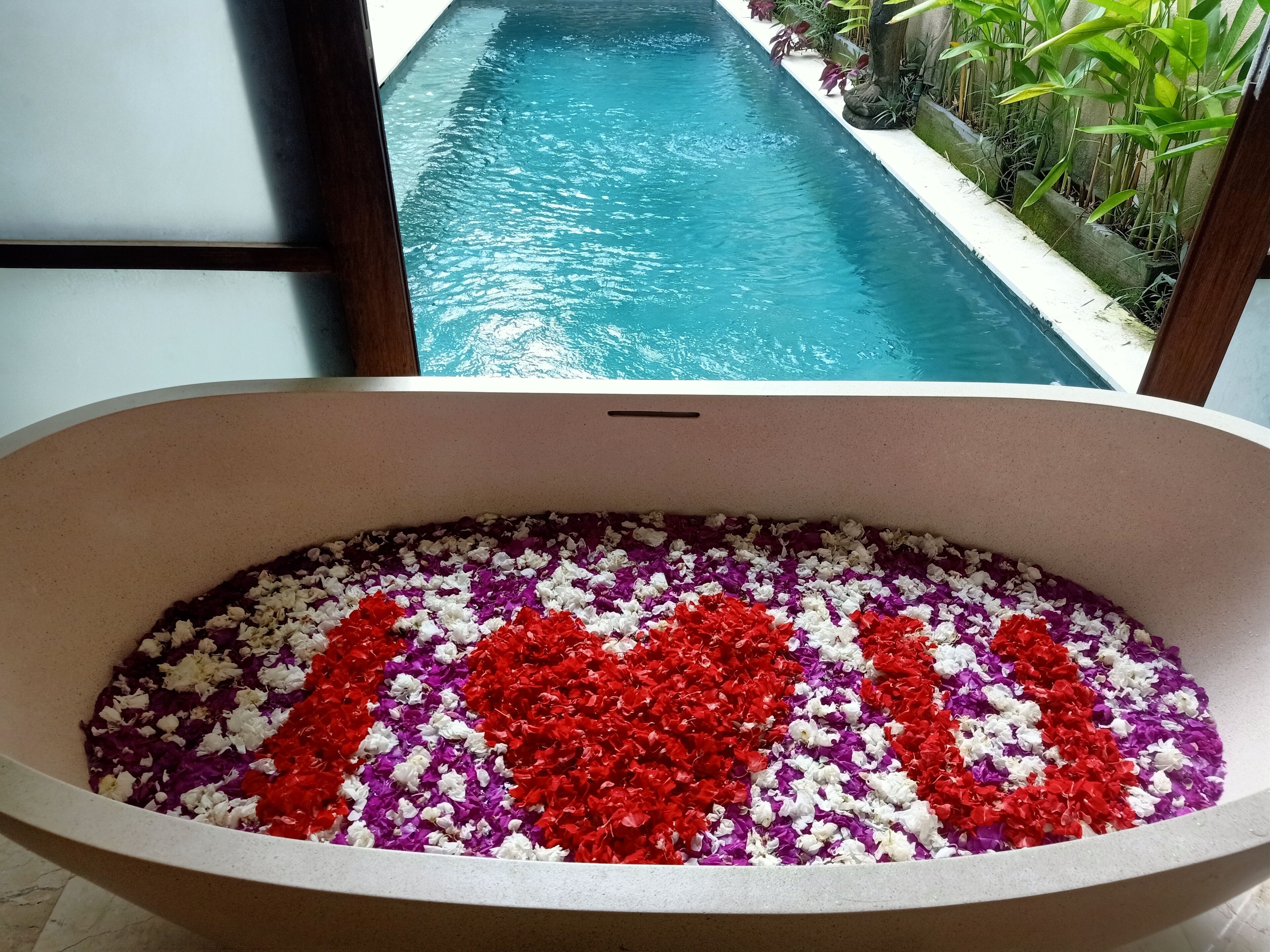Una bañera llena de pétalos de flores de colores, una vista de una piscina y un atisbo de vegetación al exterior.