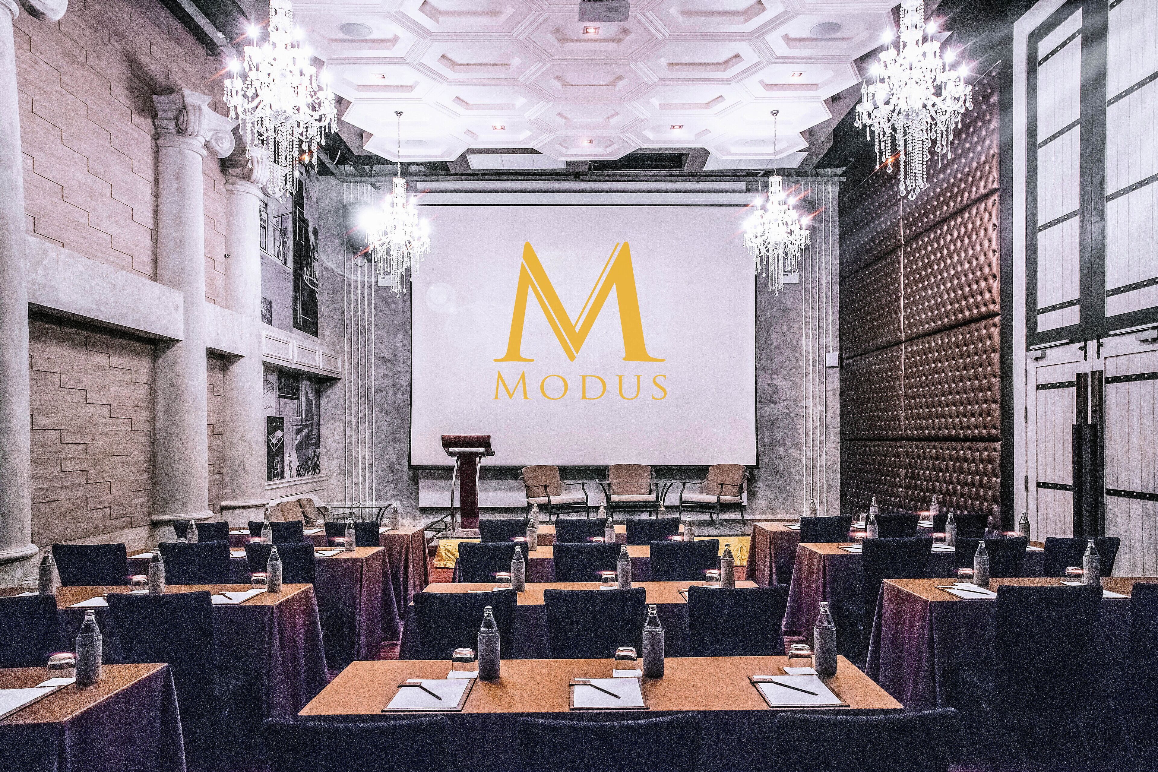Una sala conferenze con un grande schermo che mostra il logo “MODUS” in lettere dorate, circondata da tavoli con sedie, bottiglie d'acqua e blocchi per appunti.