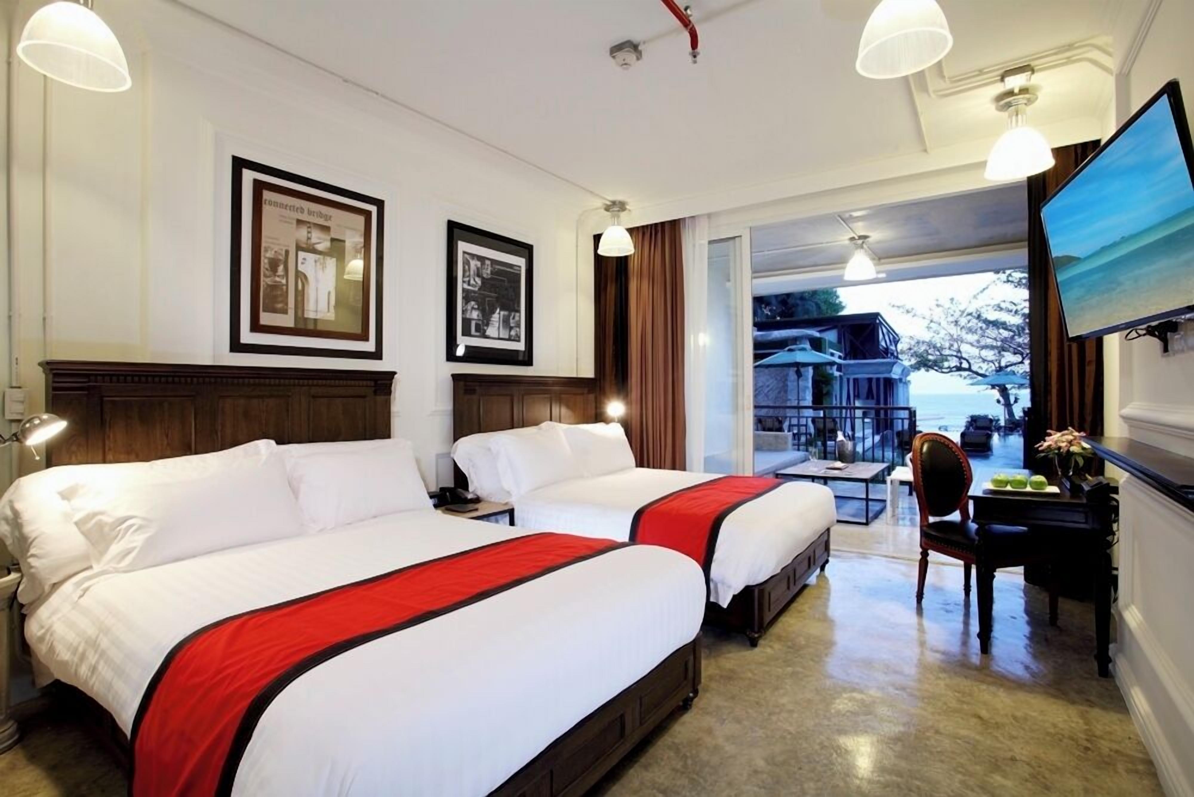 A hotel room with two beds, a desk, a chair, and a TV. There are framed pictures on the wall and a view of the outside.