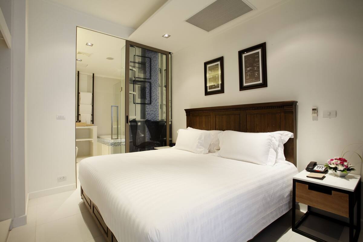A hotel room with a large bed, a wooden headboard, a bedside table with a phone and a vase of flowers, and a glass door leading to a bathroom.