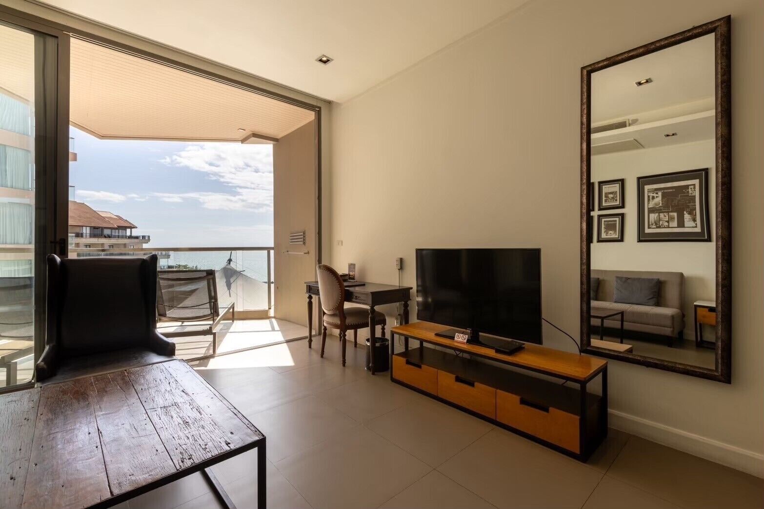 A modern living room with a TV, wooden furniture, a balcony with a view, and a large mirror.