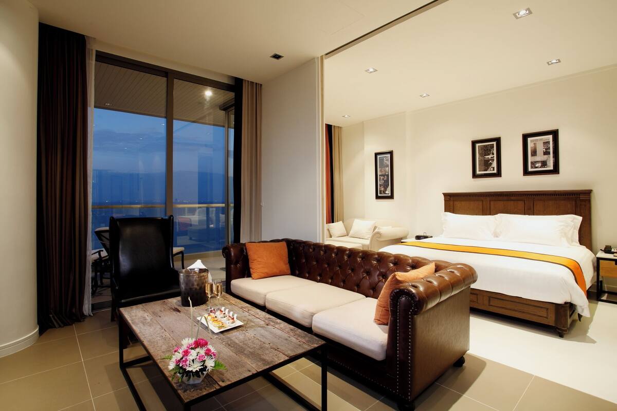 A hotel room with a large bed, a sofa, a dining table with flowers, and a balcony with a view.