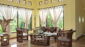 Una habitación con una mesa redonda de vidrio rodeada de sillas de mimbre, ventanas amplias con cortinas de gasa y una escultura decorativa de árbol.