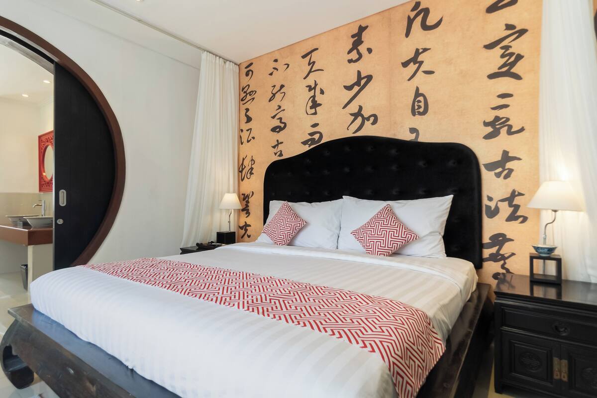 A neatly made bed with a patterned bedspread, a dark upholstered headboard, and decorative pillows. A wooden panel with calligraphy decorates the wall behind the bed.