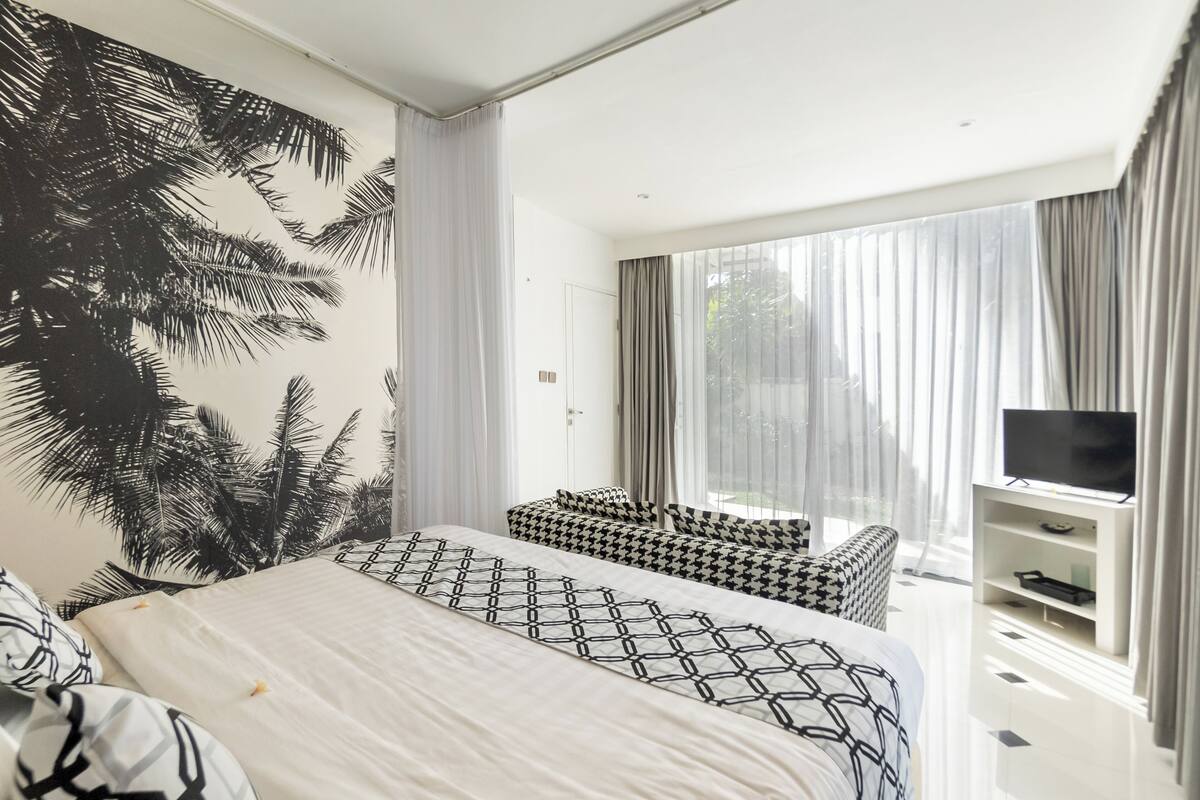 A modern hotel room with a large bed, a sofa, a TV, and a large window with curtains.