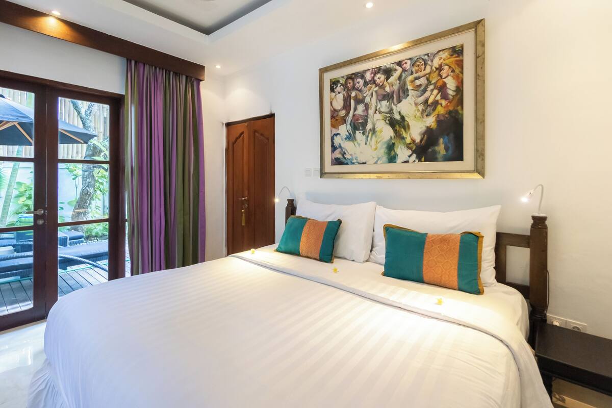 A hotel room with a large bed, a painting on the wall, and a view of the outdoors through a sliding door.