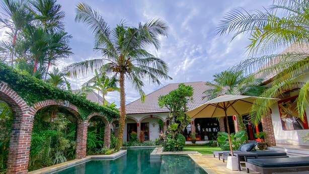 A pool area with a brick archway, palm trees, and lounge chairs. - Dyana Villas (Seminyak)