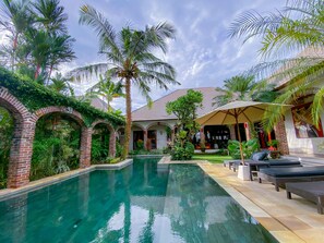 A pool area with a brick archway, palm trees, and lounge chairs. - Dyana Villas (Seminyak)